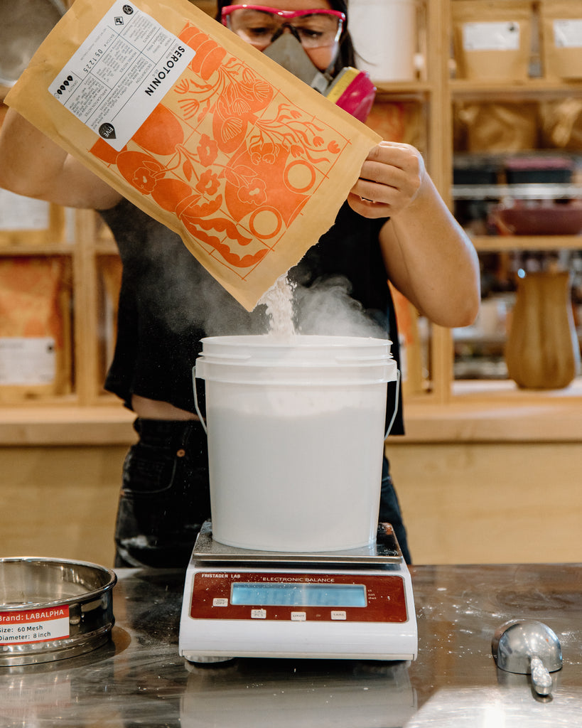 Person pouring a bag of dry glaze into a white bucket on a digital scale in a studio setting.